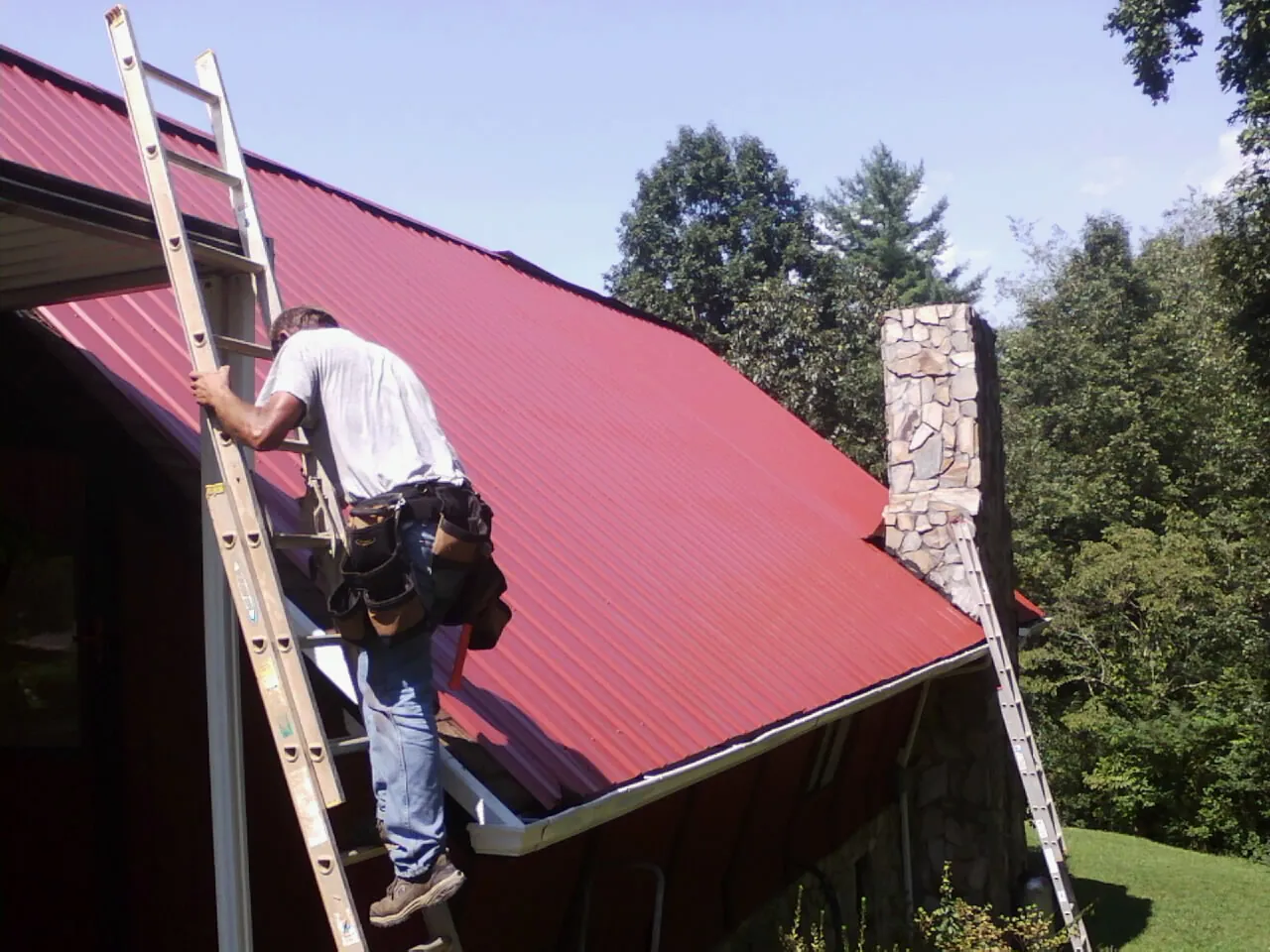 Roofer on a ladder beside a red metal roof and stone chimney — Soffit & Fascia Repair by Roofing Brothers in Saint Leonard
